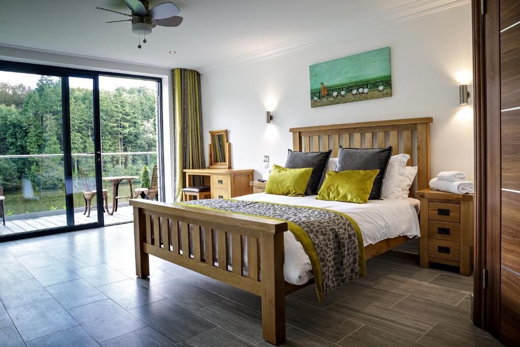 Een slaapkamer met een houten bed, een balkon met uitzicht op groen, een schilderij aan de muur en een plafondventilator.