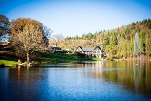 Un albergue junto al lago con un edificio grande, un área verde y una colina boscosa al fondo.