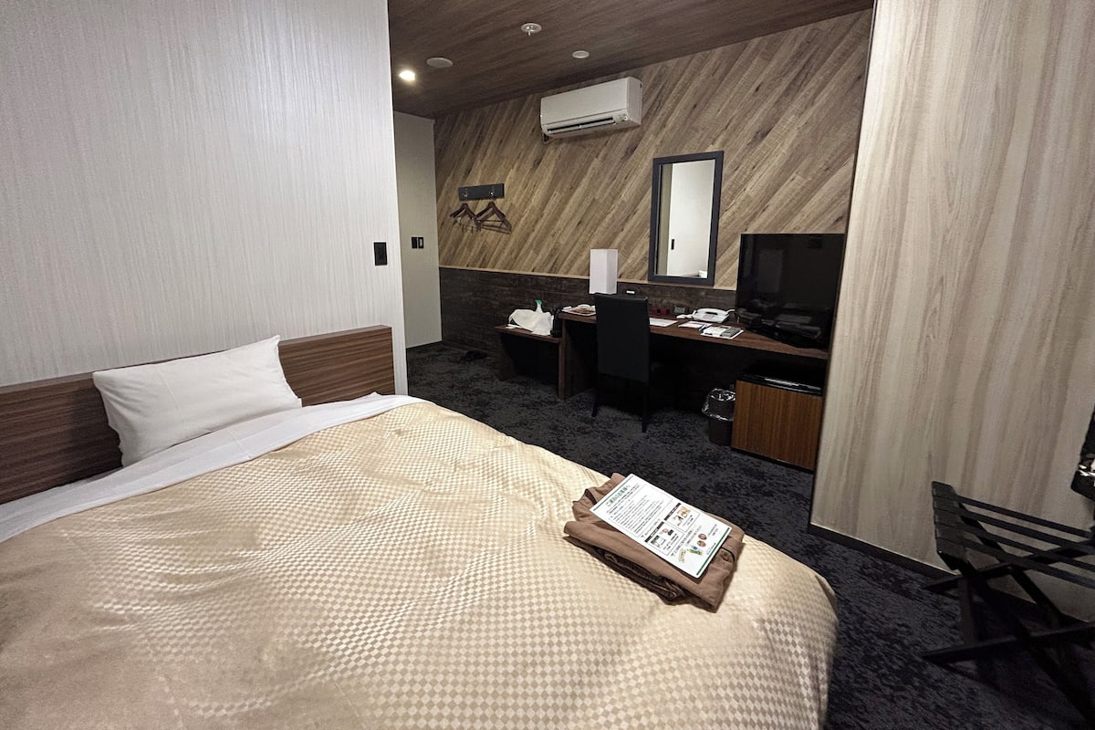 A hotel room with a bed, a desk, a chair, an air conditioning unit, and a mirror.