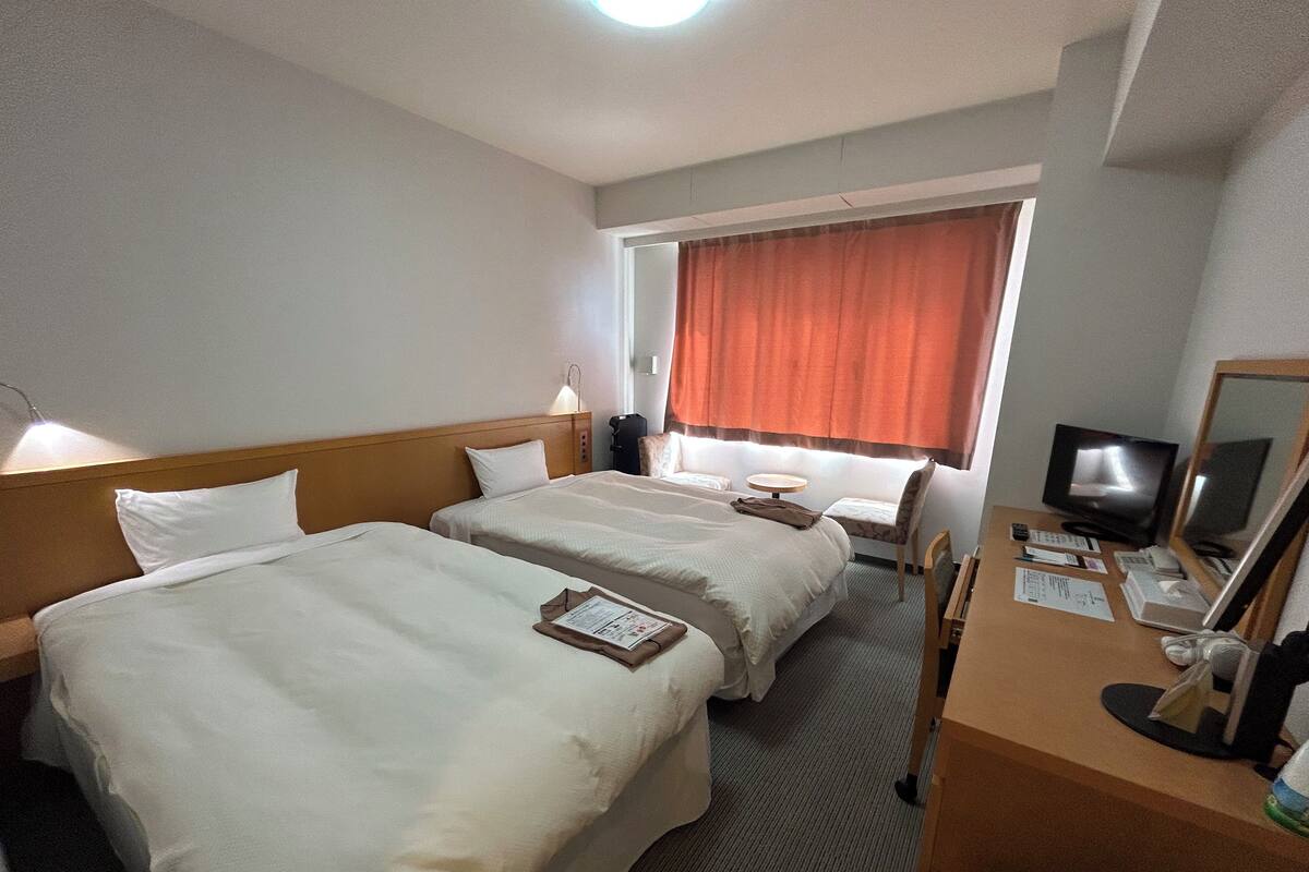 A hotel room with two beds, a desk with a computer, a TV, and a window with curtains.