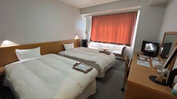 Ein Hotelzimmer mit zwei Betten, einem Schreibtisch mit Computer, einem Fernseher und einem Fenster mit Vorhängen.