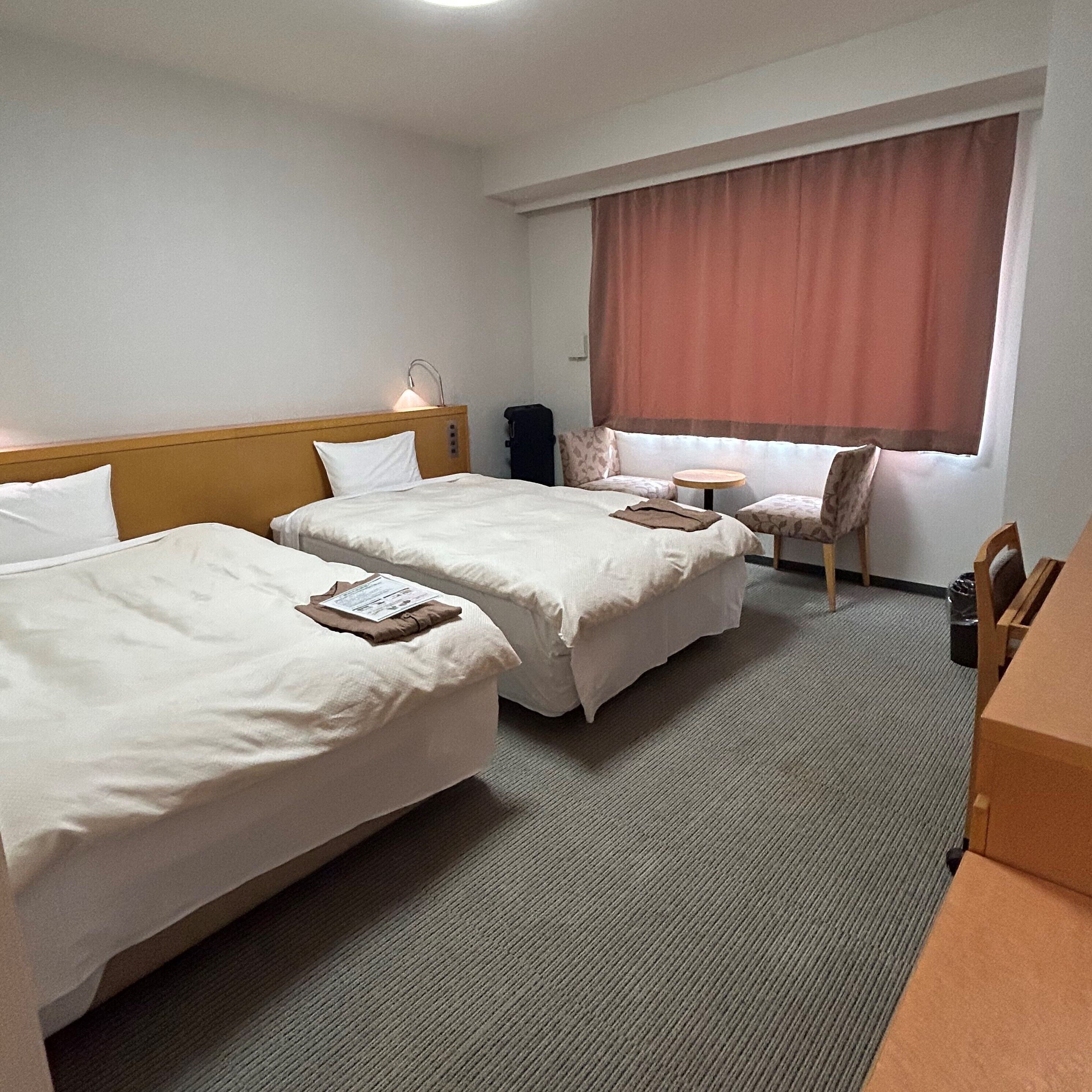 A hotel room with two single beds, a small table, a chair, and a window with curtains.