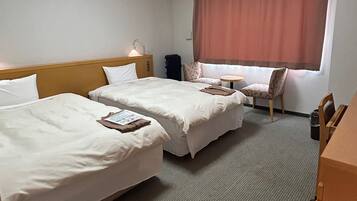 A hotel room with two single beds, a small table, a chair, and a window with curtains.