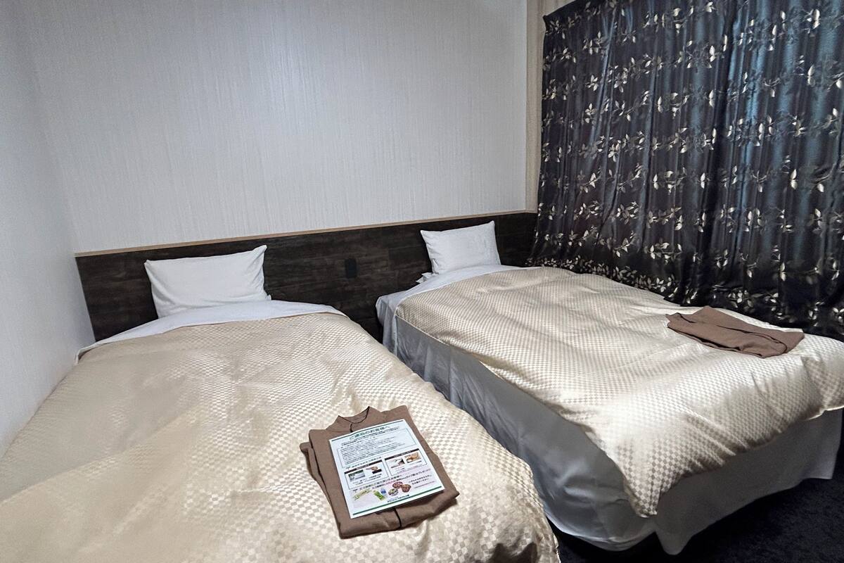 A hotel room with two beds, each with a beige bedspread and white pillows. A brown jacket is folded on the right bed. The room has a dark curtain with a floral pattern on the right side.