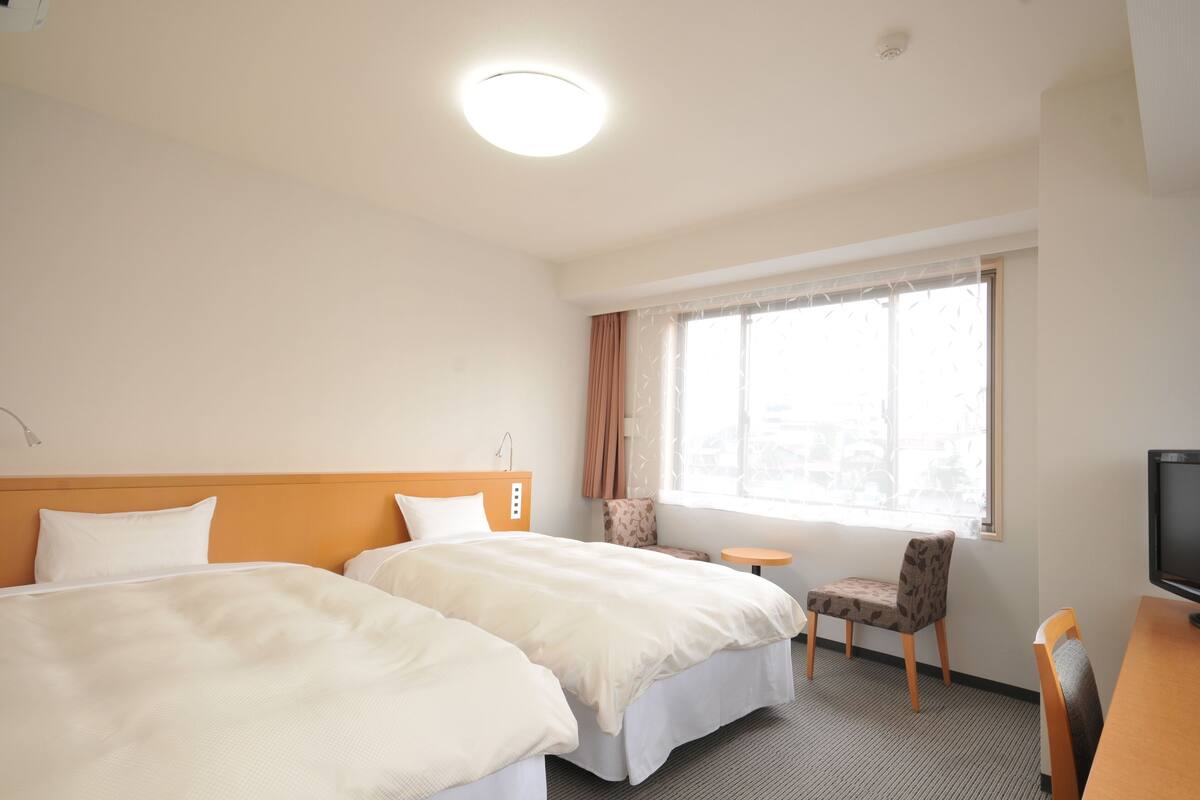 A hotel room with two beds, a TV, a small table, and a window with curtains.
