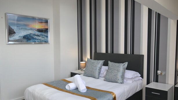 A neatly made bed with a striped wall behind it, a bedside table with a lamp, and a framed picture on the wall. - Hotel 33 (Blackpool)