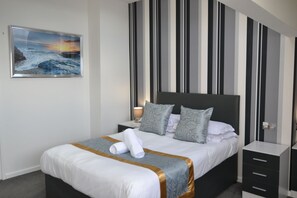 A neatly made bed with a striped wall behind it, a bedside table with a lamp, and a framed picture on the wall. - Hotel 33 (Blackpool)