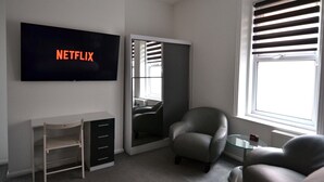 A modern room with a TV displaying 'NETFLIX', a desk with drawers, a chair, a glass table, and a window with blinds. - Hotel 33 (Blackpool)