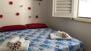 Ein Schlafzimmer mit einem Bett, roten Kissen, einer blumigen Tagesdecke, einem Gemälde mit Strandmotiv und einem Fenster mit Jalousien.
