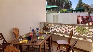 Ein Holztisch mit Stühlen auf einem Balkon, ein Gitterzaun und im Hintergrund eine Palmenlandschaft sowie ein Gebäude.