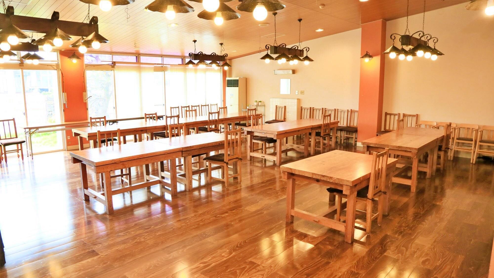 A spacious dining area with multiple wooden tables and chairs, large windows, and hanging pendant lights.