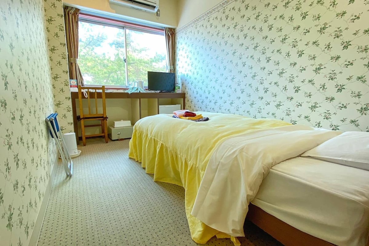 Une chambre à coucher avec un lit, un bureau avec un ordinateur, une chaise et une fenêtre.