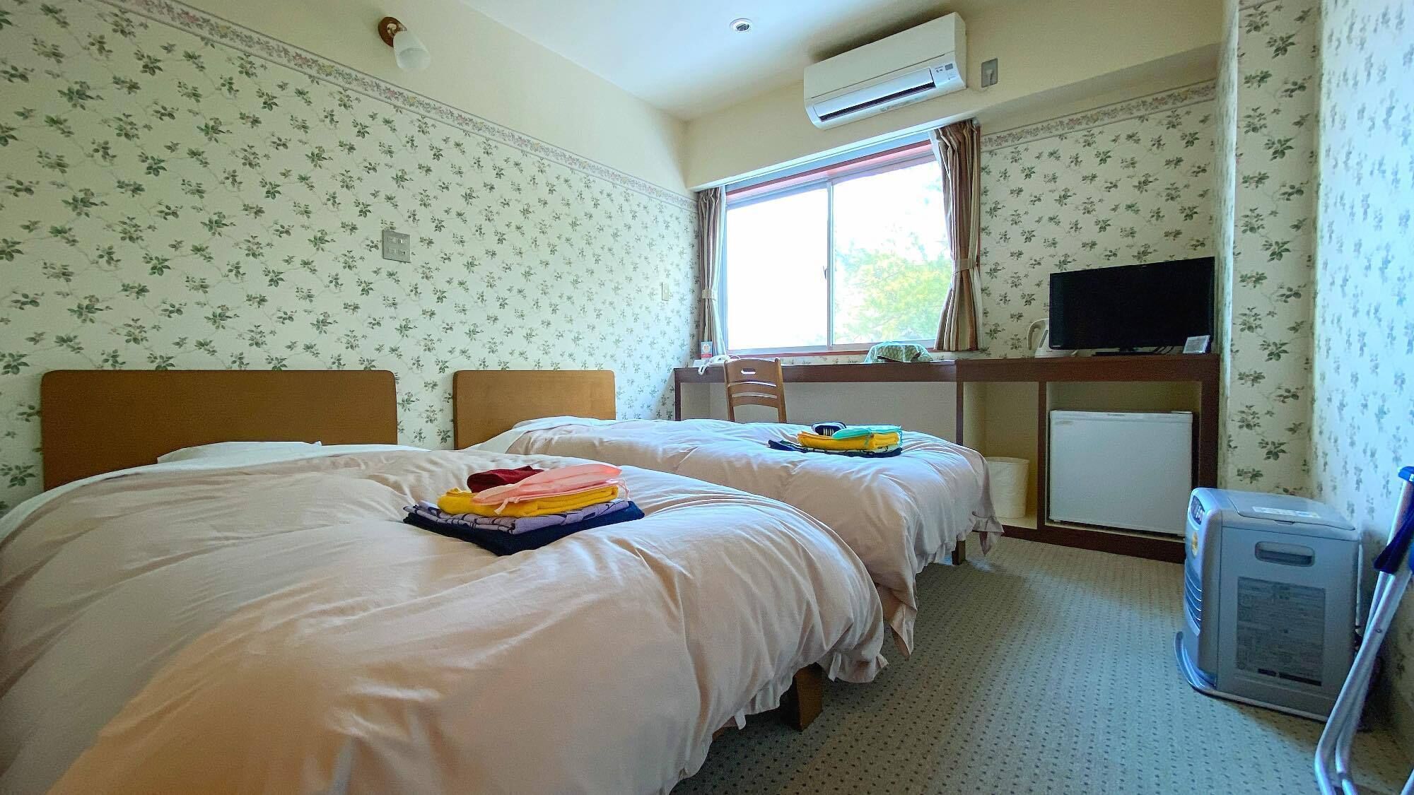 A hotel room with two beds, a desk with a computer, an air conditioner, and a window with curtains.
