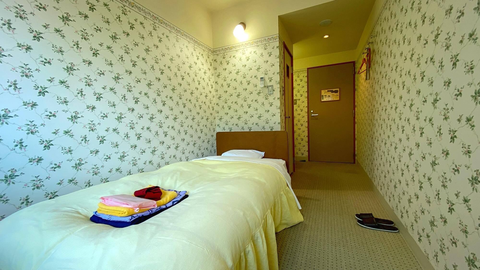 A hotel room with floral wallpaper, a bed with a white headboard, a bedside table with a lamp, and a door leading to another room.