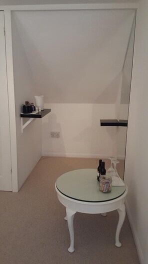 Una habitación pequeña y blanca con una mesa redonda de vidrio, una puerta blanca y una pared blanca. Hay una estantería con una cafetera y tazas, además de una botella y un frasco sobre la mesa.