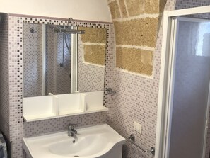 A bathroom with a tiled wall, a sink, a mirror, and a shower area.