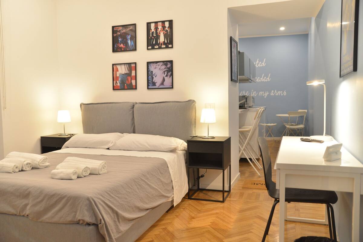 A hotel room with a bed, bedside tables, a desk, and chairs. There are wall art pieces and a wall-mounted TV.