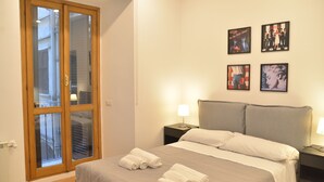 A hotel room with a bed, two bedside tables, a nightstand, a window with blinds, and a door with a glass panel. - All You Need Is Rome Guesthouse (Rome)