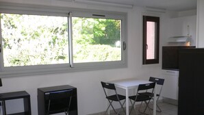 Une petite cuisine avec une table et des chaises blanches, un bureau noir et une fenêtre donnant sur un paysage verdoyant.