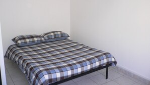 Une petite chambre avec un lit simple, une couvre-lit et des coussins à carreaux, des murs blancs et un sol carrelé.