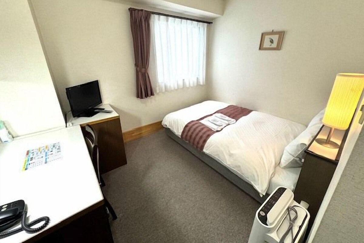 Habitación de hotel con cama, escritorio, computadora, teléfono y ventana con cortinas.