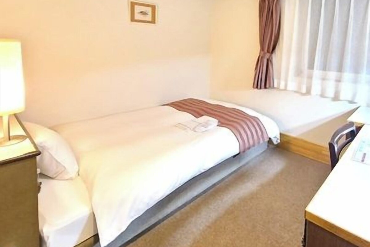 A small, single-bed room with a wooden headboard, a bedside table with a lamp, and a window with curtains.