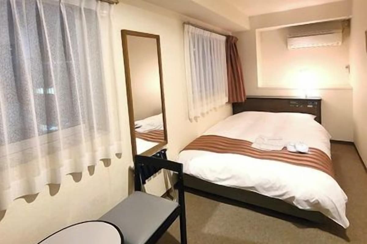 A hotel room with a bed, a chair, a desk, a mirror, and curtains.