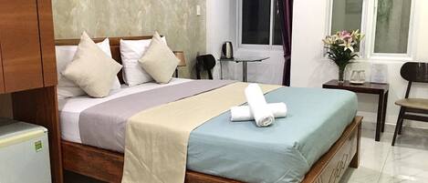 Habitación de hotel con cama de madera, un escritorio pequeño, una silla, ventana con cortinas y un refrigerador.