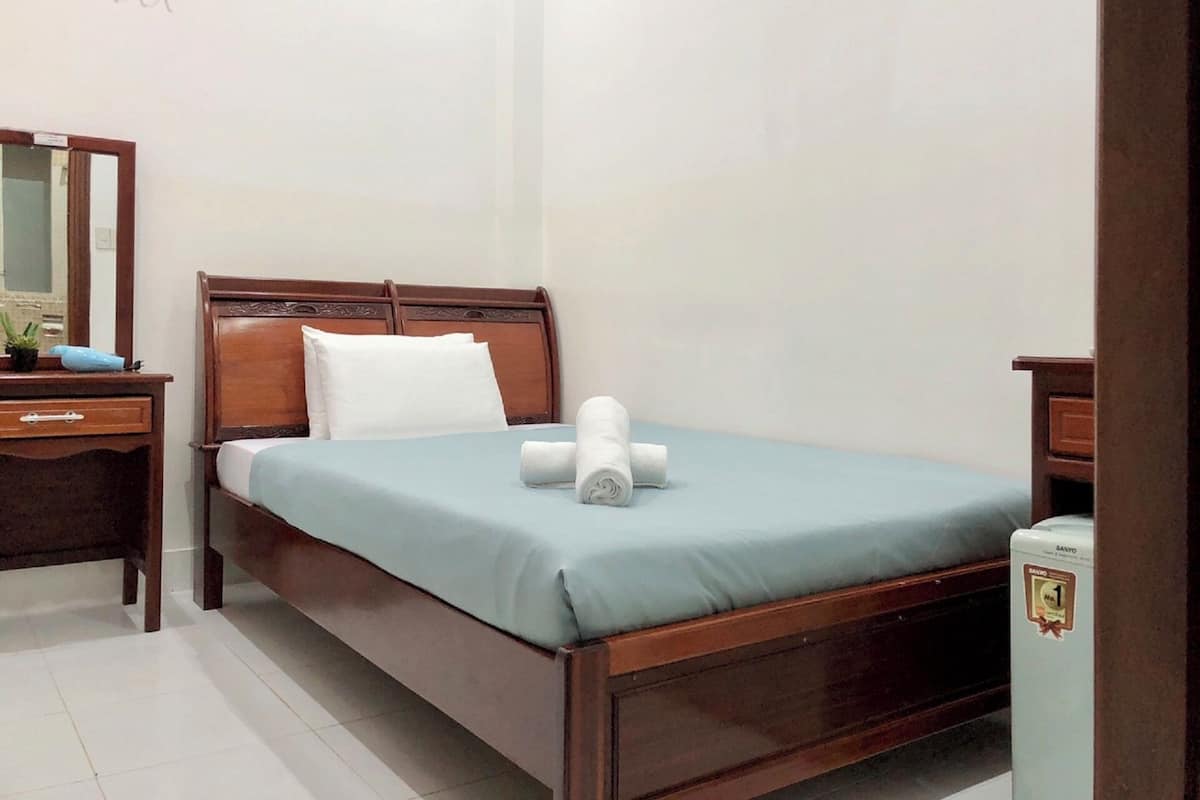 Un lit bien fait, recouvert d’une couvre-lit bleue et agrémenté d’oreillers blancs, une table de chevet en bois avec une lampe, et une petite table d’appoint avec un miroir.