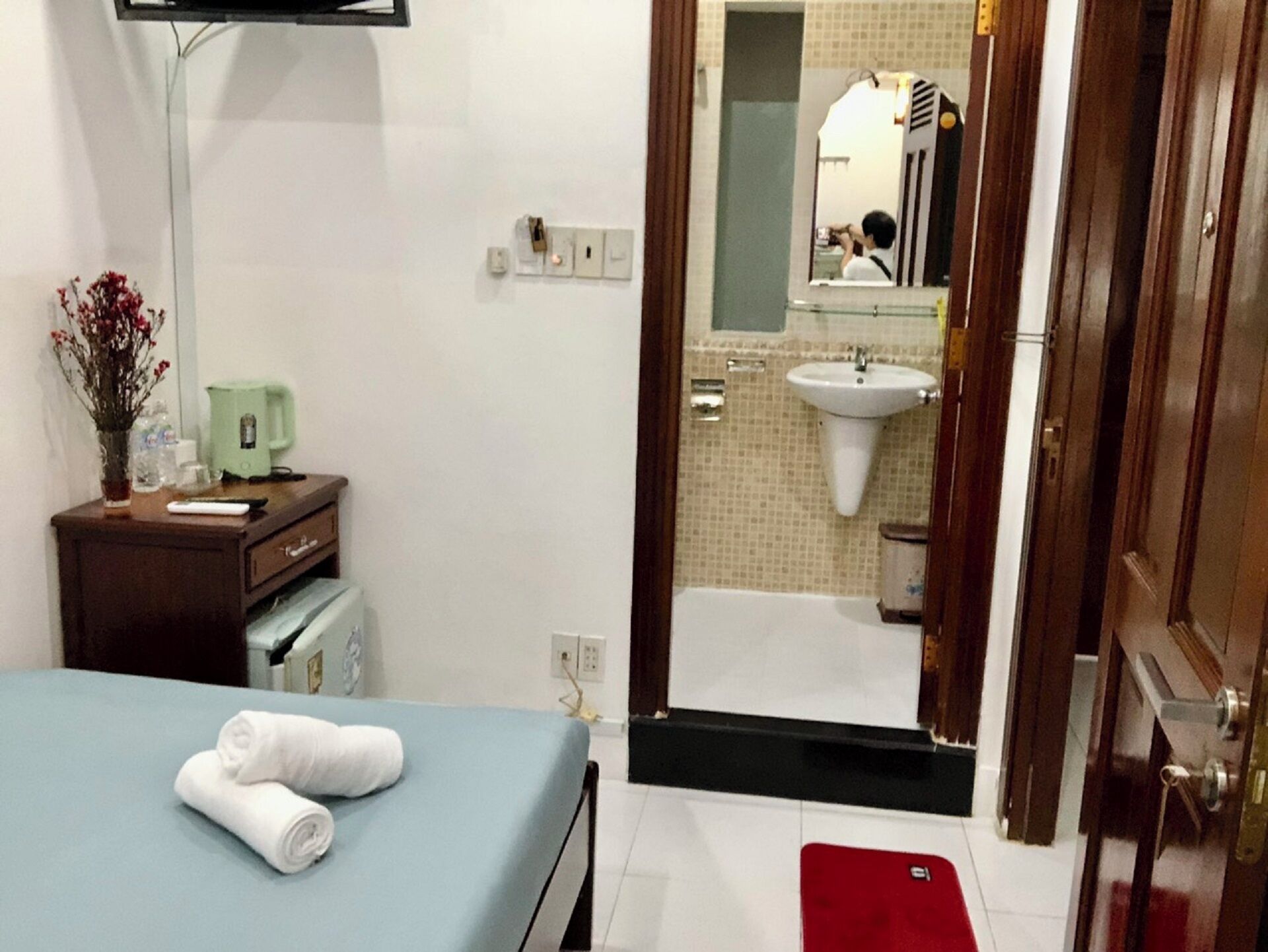 Habitación de hotel con una cama, un escritorio con lámpara, cafetera y baño visible a través de una puerta abierta.
