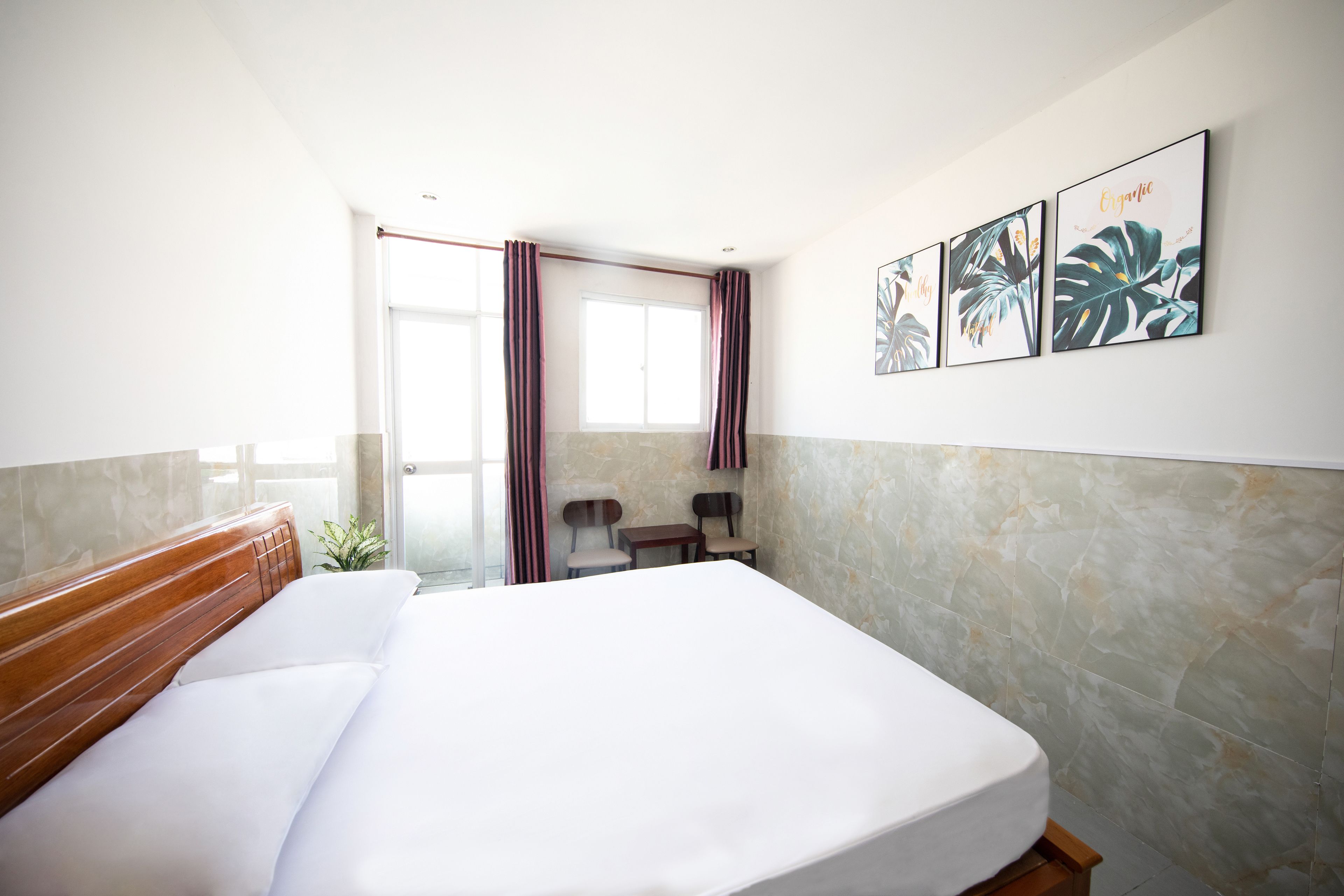 Ein Hotelzimmer mit einem großen Bett, einem kleinen Tisch, zwei Stühlen, einem Fenster mit Vorhängen und drei gerahmten Bildern an der Wand.