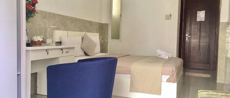 A modern hotel room with a bed, a desk with a chair, a small table with a vase of flowers, an air conditioning unit, and a door.