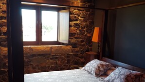 A bedroom with a stone wall, a bed with patterned bedding, a window with shutters, and a nightstand with a lamp. - Casona Los Gamonales (Carreno)