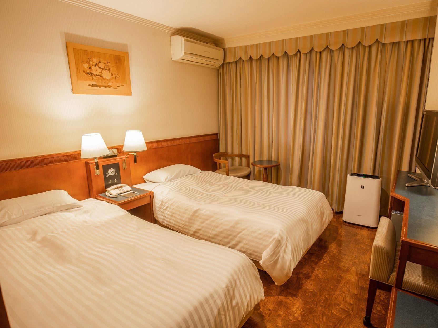 A hotel room with two beds, a desk, a chair, a television, an air conditioning unit, and a painting on the wall.