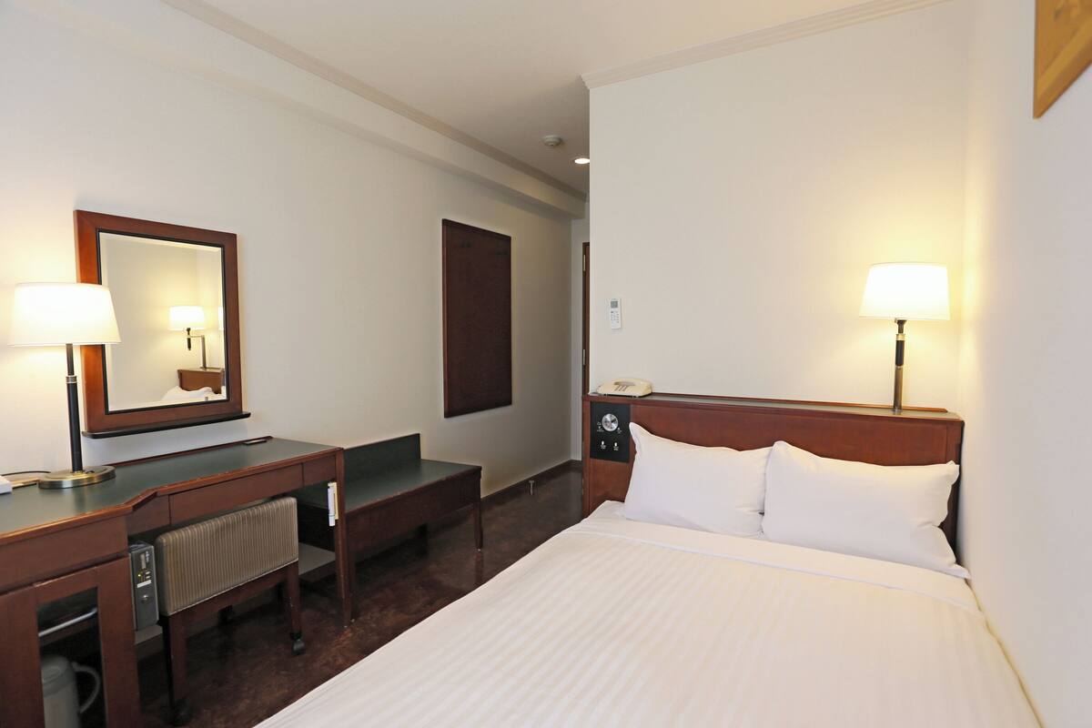 Een hotelkamer met een bed, nachtkastje, lamp, bureau, stoel en een spiegel.