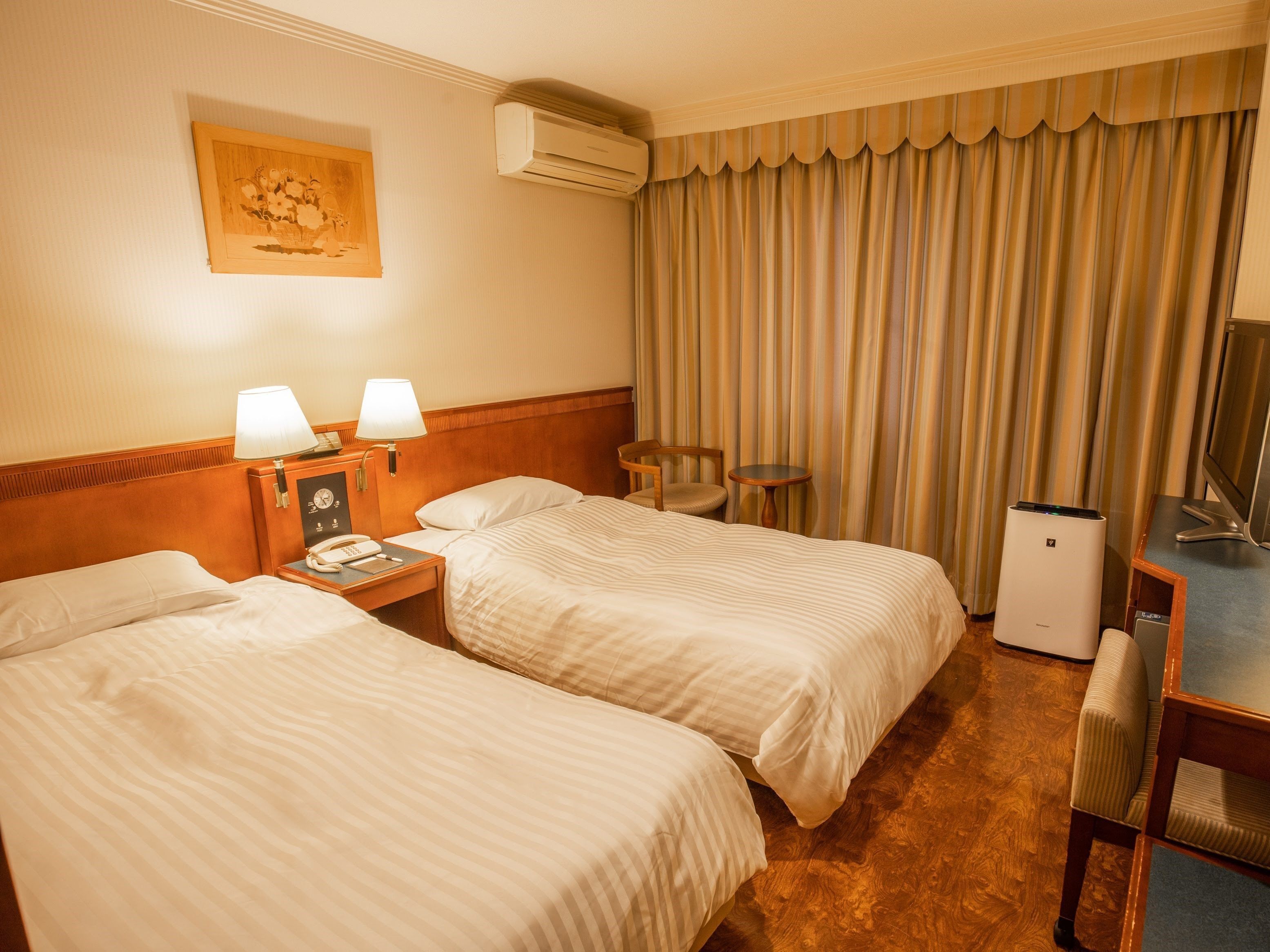 A hotel room with two beds, a desk with a computer, a chair, a lamp, a clock, and an air conditioning unit.