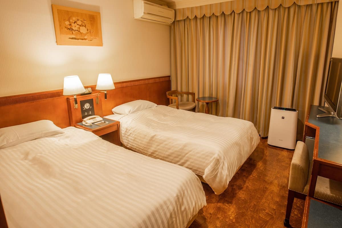 Een hotelkamer met twee bedden, een bureau met een computer, een stoel, een lamp, een klok en een airconditioning.