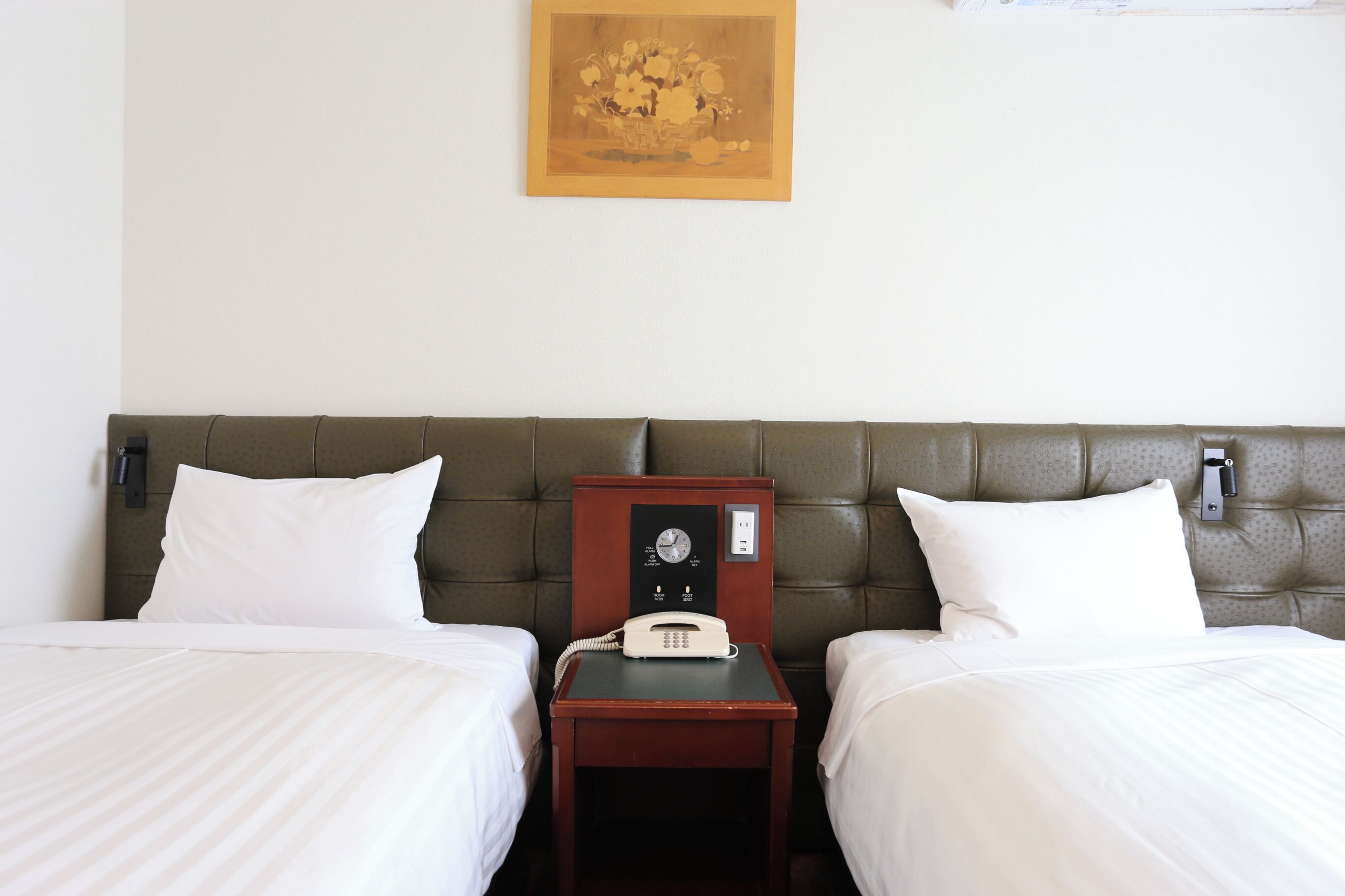 Ein Hotelzimmer mit zwei Betten, einem Nachttisch aus Holz mit Telefon und einem an der Wand montierten Lichtschalter.