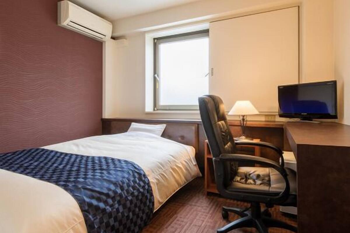 A hotel room with a bed, a desk with a chair, a television, and an air conditioning unit.