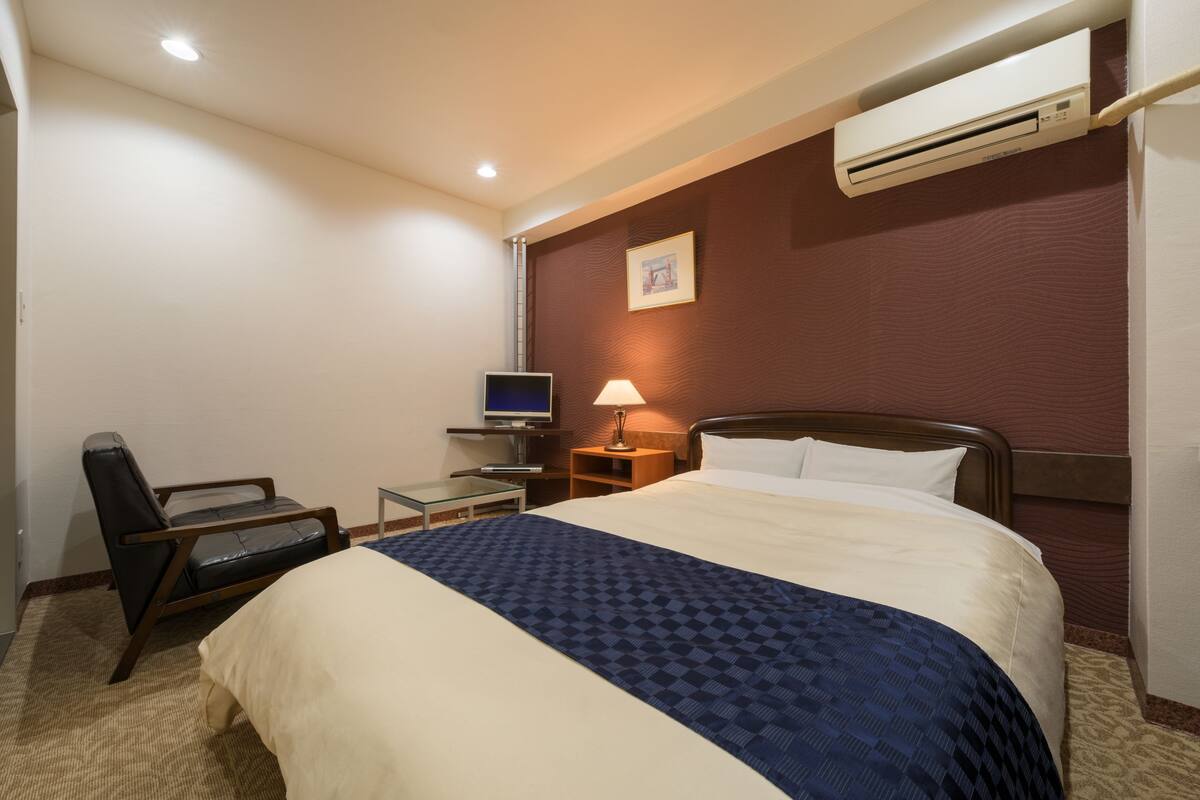 A hotel room with a bed, a desk with a computer, a chair, a lamp, and an air conditioner.