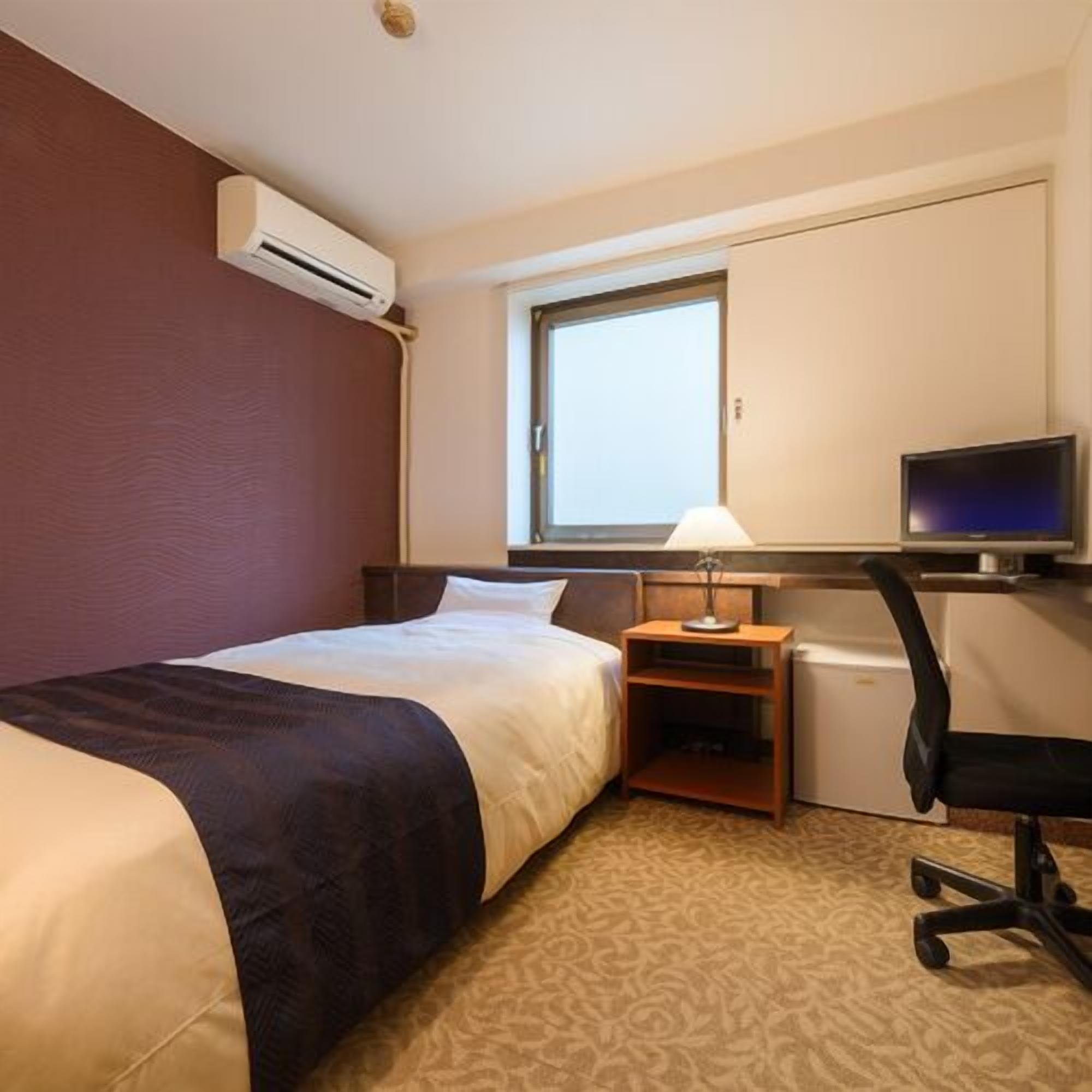 Una habitación de hotel con cama, escritorio con computadora, silla, lámpara y equipo de aire acondicionado.