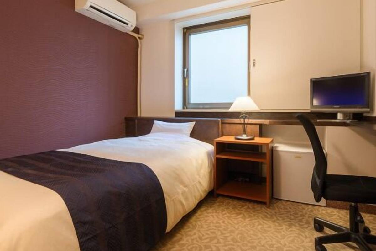 A hotel room with a bed, a desk with a computer, a chair, a lamp, and an air conditioning unit.