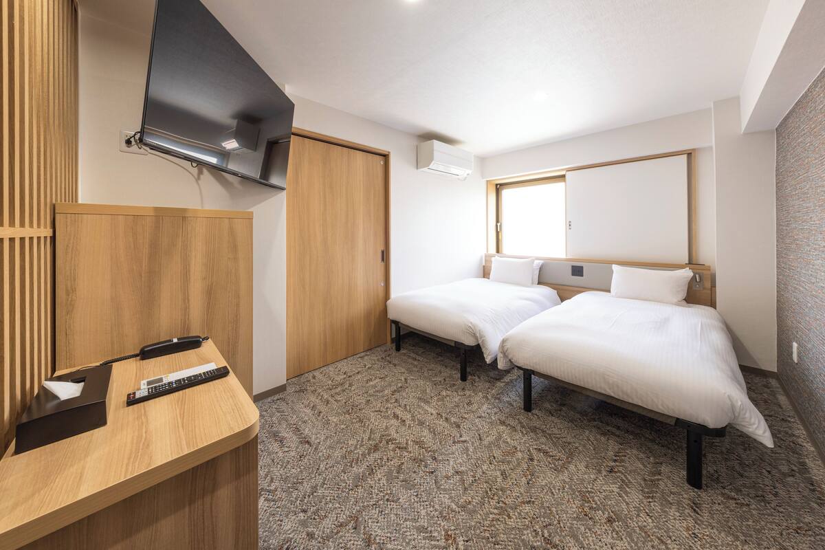 A hotel room with two beds, a wooden desk with a telephone and remote control, a flat-screen TV mounted on the wall, and a window with a view of the outside.