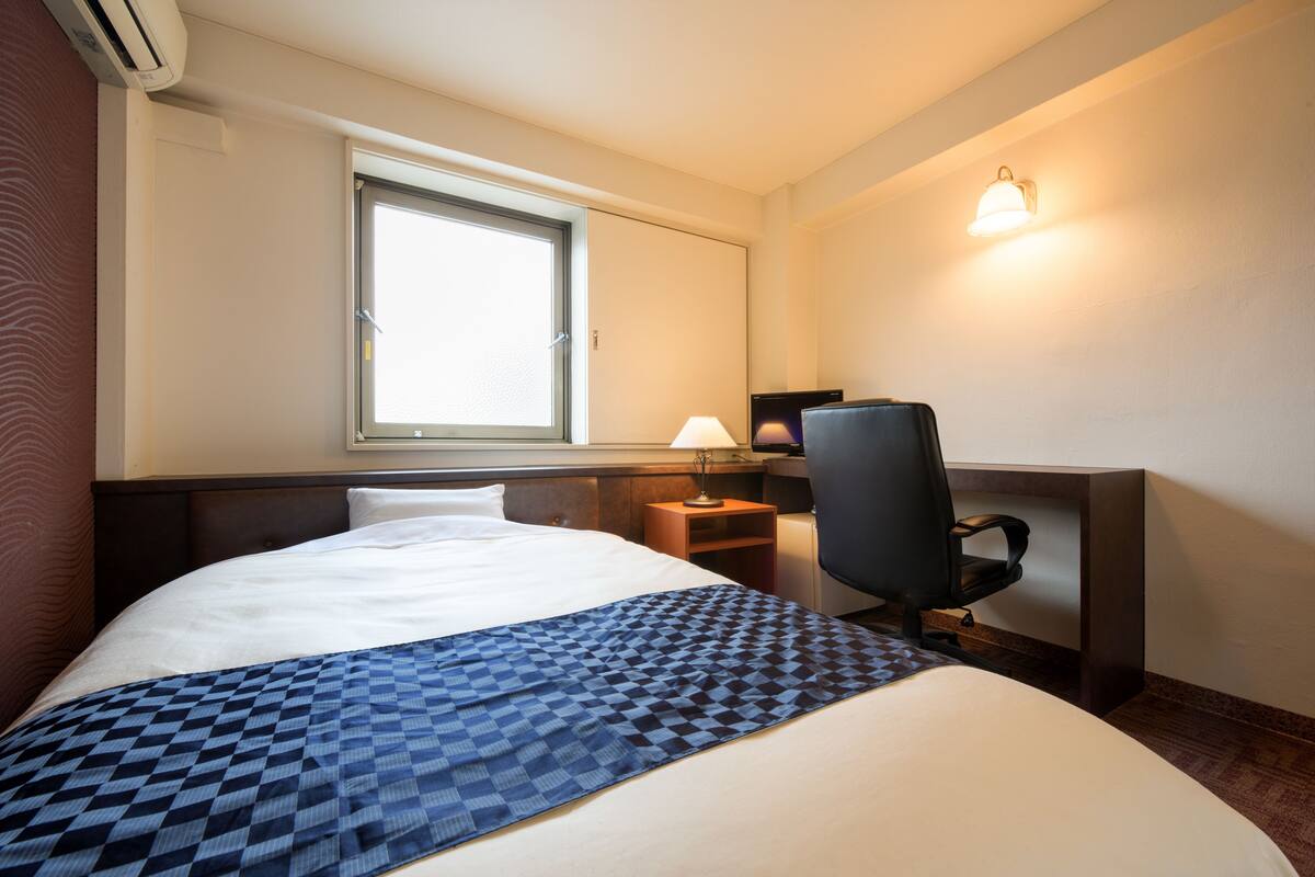 A hotel room with a large bed, a desk with a chair, a lamp, and a window.