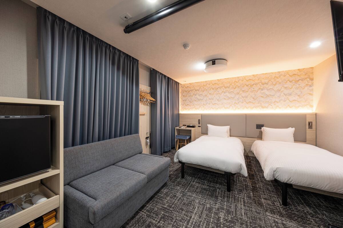 A hotel room with a grey sofa, two single beds, a TV, and a desk with a chair.