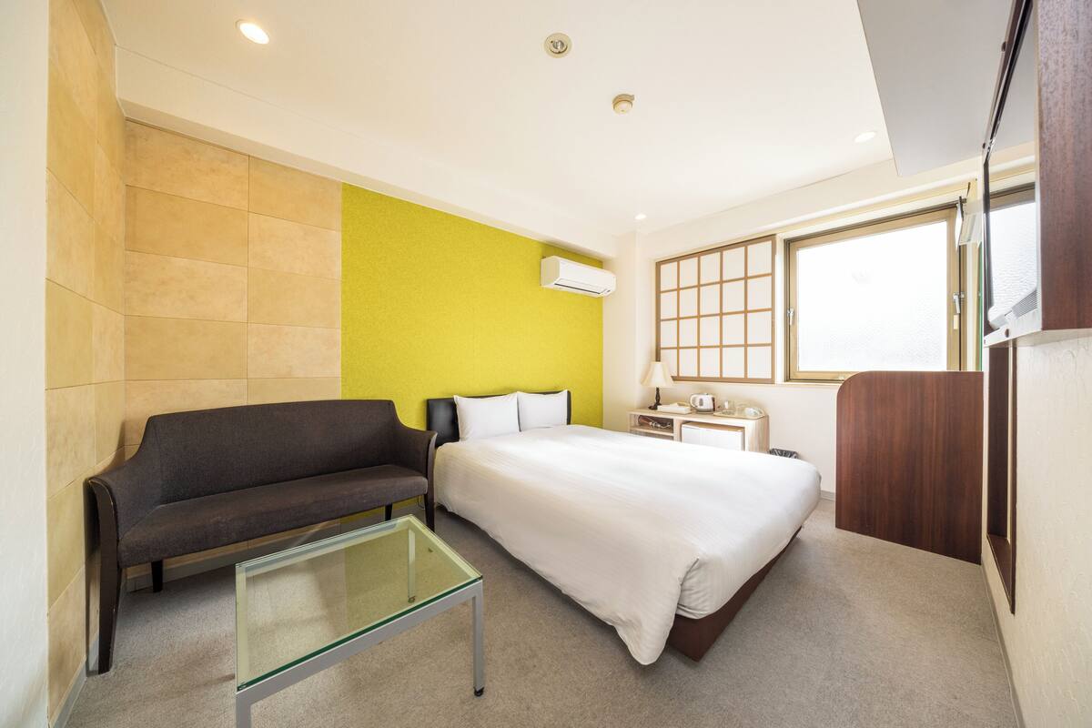 A hotel room with a bed, a sofa, a glass coffee table, a nightstand, and a window with a view.