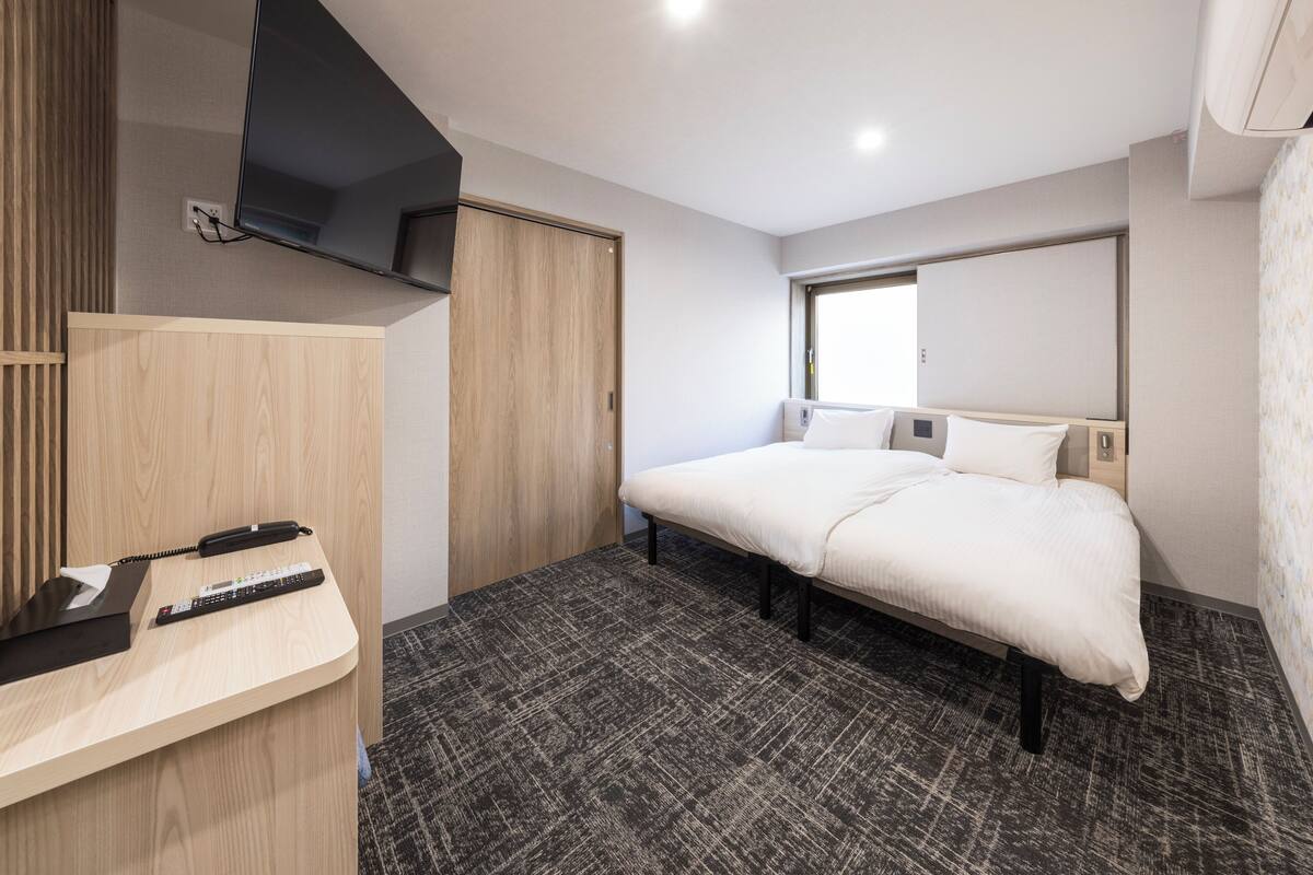 A modern hotel room with a bed, a desk with a telephone and remote control, a wardrobe, and a large window.