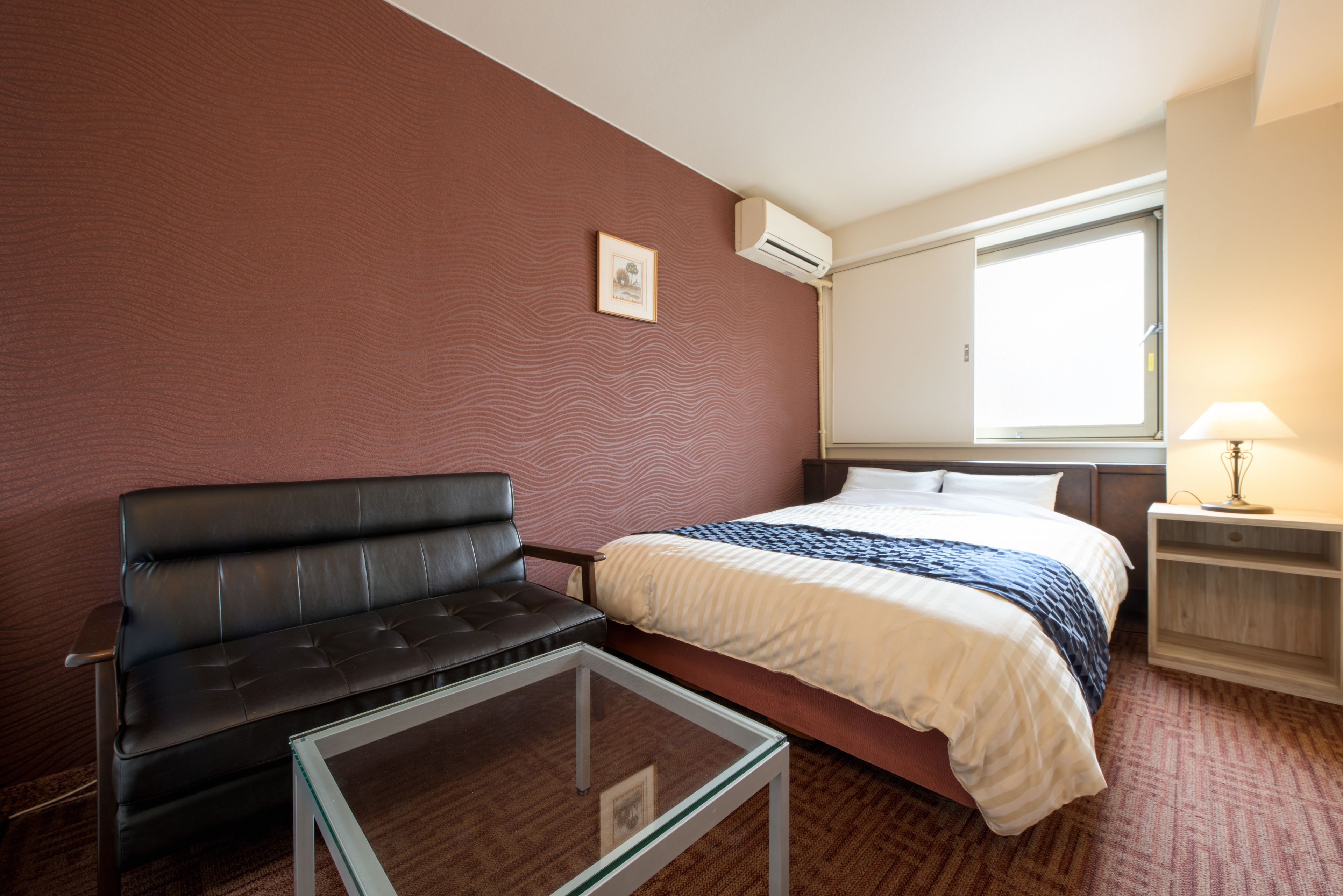 A hotel room with a bed, a sofa, a glass-top table, a nightstand with a lamp, and a window with blinds.