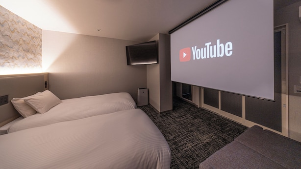 Habitación de hotel con dos camas, un televisor de pantalla plana montado en la pared y una pantalla de proyección que muestra el logotipo de YouTube.