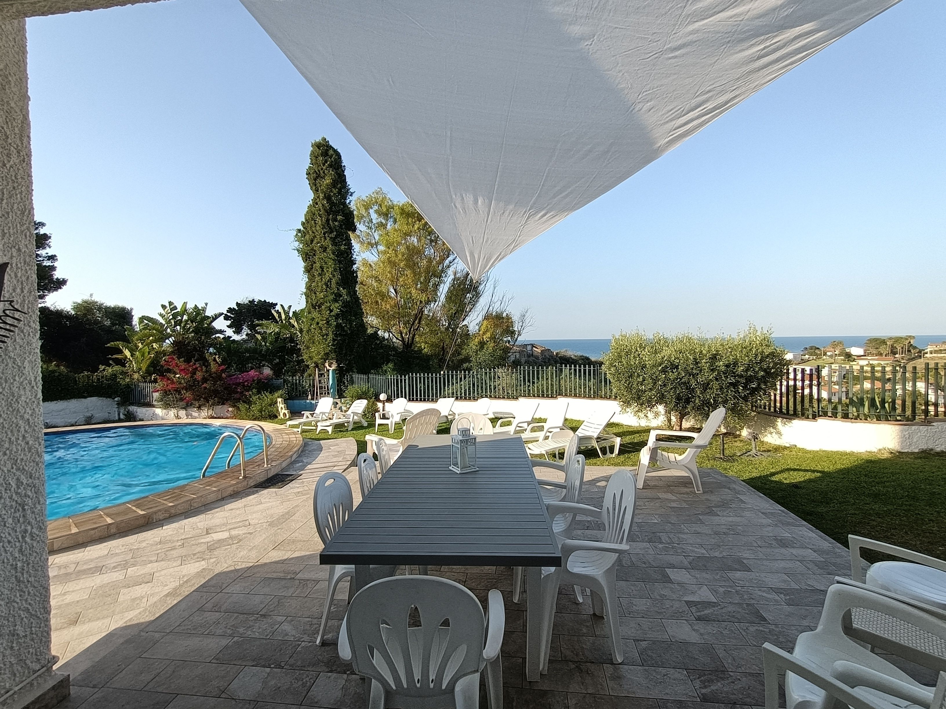 An outdoor pool area with a table and chairs, surrounded by a white fence and greenery.